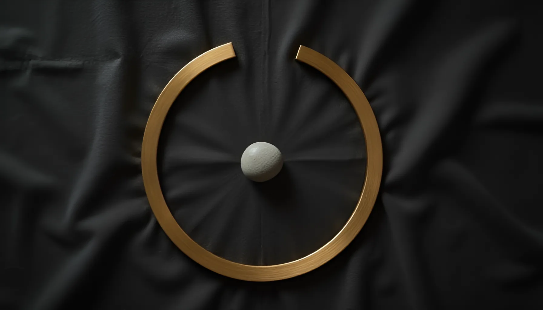 어두운 천 위에 놓인 한쪽이 트인 황동빛 고리와 그 안쪽 가운데의 옅은 회색 조약돌